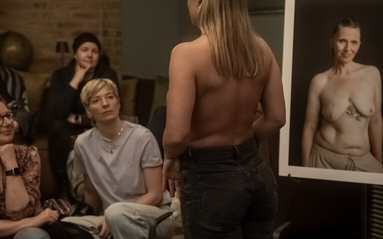 Eine Frau zeigt ihre Brustkrebsnarbe. Rundherum stehen Bilder von Frauen, die ihre Narben fotografieren ließen. | © Foto: Lana Immelmann