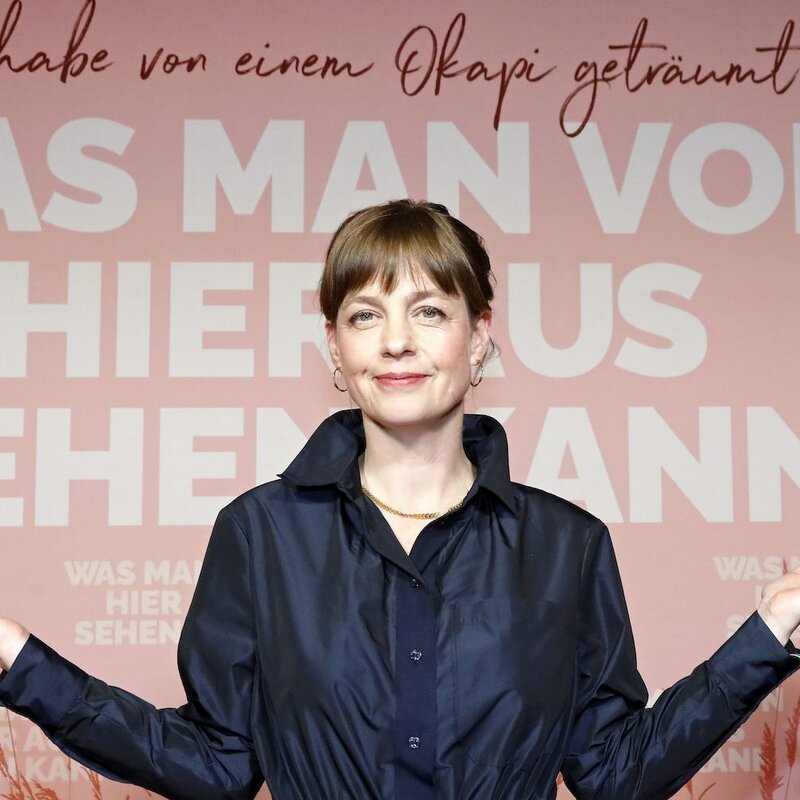 Bestsellerautorin Mariana Leky steht vor dem Filmplakat von „Was man von hier aus sehen kann“, darauf abgebildet ist ein Okapi und der Titel, der Hintergrund ist rosa, Mariana trägt einen dunkelblauen Mantel und richtet die Unterarme nach rechts und links aus. | © „Was man von hier aus sehen kann“ Weltpremiere 
ASTOR Film Lounge im ARRI, München 
©Kurt Krieger/STUDIOCANAL