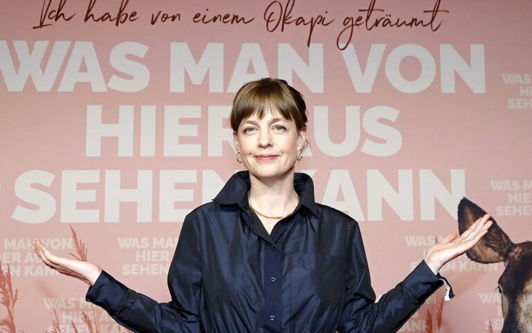 Bestsellerautorin Mariana Leky steht vor dem Filmplakat von „Was man von hier aus sehen kann“, darauf abgebildet ist ein Okapi und der Titel, der Hintergrund ist rosa, Mariana trägt einen dunkelblauen Mantel und richtet die Unterarme nach rechts und links aus. | © „Was man von hier aus sehen kann“ Weltpremiere 
ASTOR Film Lounge im ARRI, München 
©Kurt Krieger/STUDIOCANAL