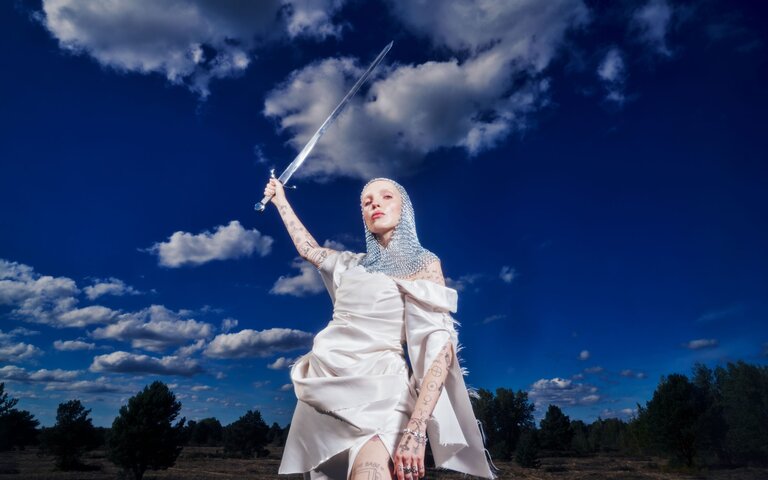Mia Morgan trägt ein weißes Kleid und eine Kettenhaube und hält ein Schwert über ihren Kopf. | © Foto: Can Wagener