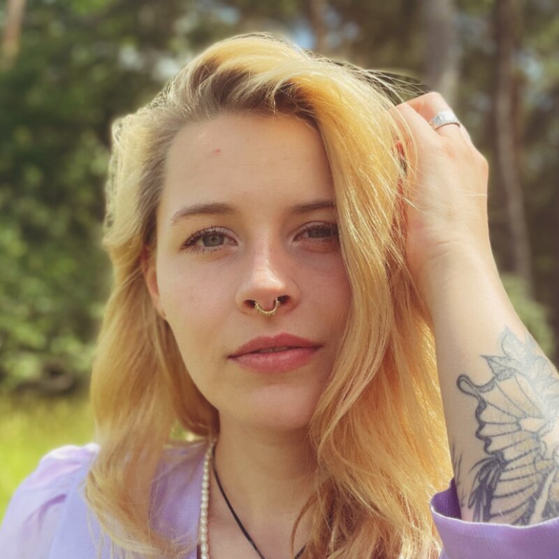 Die Autorin Carina Waldmann fasst sich mit der Linken Hand an die Haaren, trägt eine lilafarbene Bluse und blickt in die Kamera. | © Foto: David Rombach