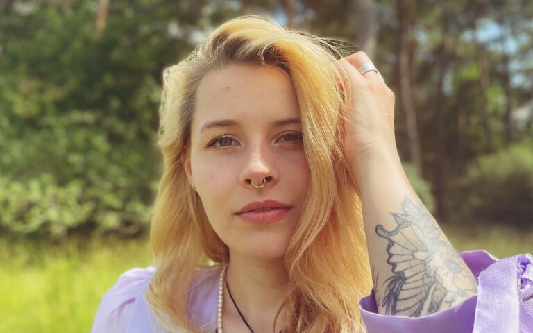 Die Autorin Carina Waldmann fasst sich mit der Linken Hand an die Haaren, trägt eine lilafarbene Bluse und blickt in die Kamera. | © Foto: David Rombach