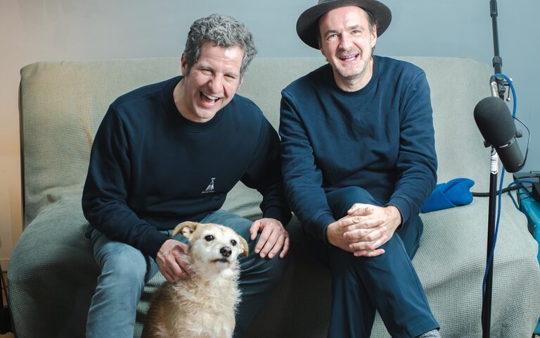 Die Podcast-Hosts Winson und Ueli sitzen im Aufnahme-Wohnzimmer, vor ihnen ein goldbrauner Hund, mittelgroß. Sie lachen in die Kamera. | © Foto: Winson