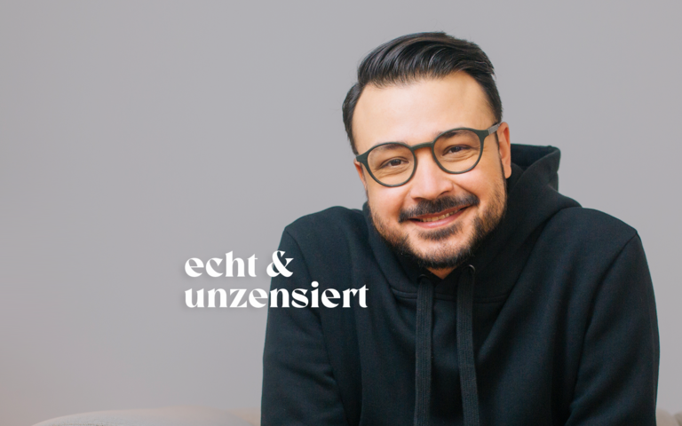 Psychologe Umut Özdemir sitzt vor einer weißen Fotowand und lächelt. | © Credit: Mehran Djojan