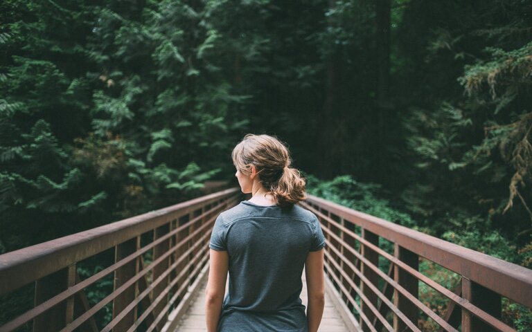 Eine Frau, von hinten zu sehen, geht über eine Brücke in Richtung Wald. | © Foto: Jake Melara/Unsplash