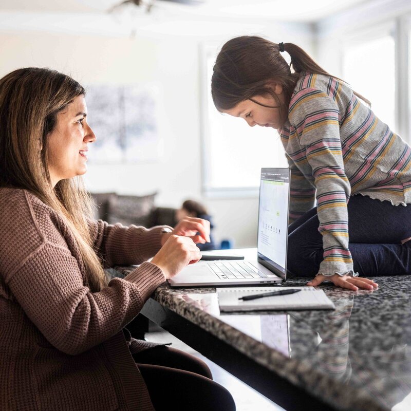 Das Bild zeigt eine am Küchentresen sitzende Frau vor ihrem Laptop. Auf dem Tresen kniet ein kleines Mädchen, das auf ihren Laptop schaut. | © Foto: Momo Productions | Getty Images 