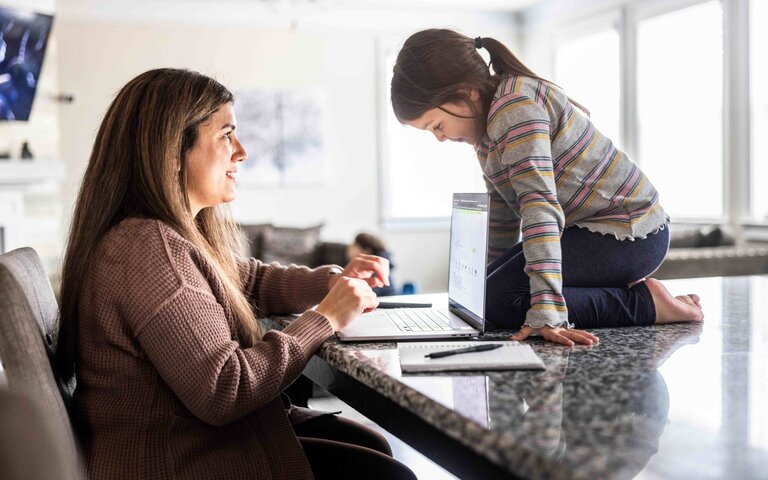 Das Bild zeigt eine am Küchentresen sitzende Frau vor ihrem Laptop. Auf dem Tresen kniet ein kleines Mädchen, das auf ihren Laptop schaut. | © Foto: Momo Productions | Getty Images 
