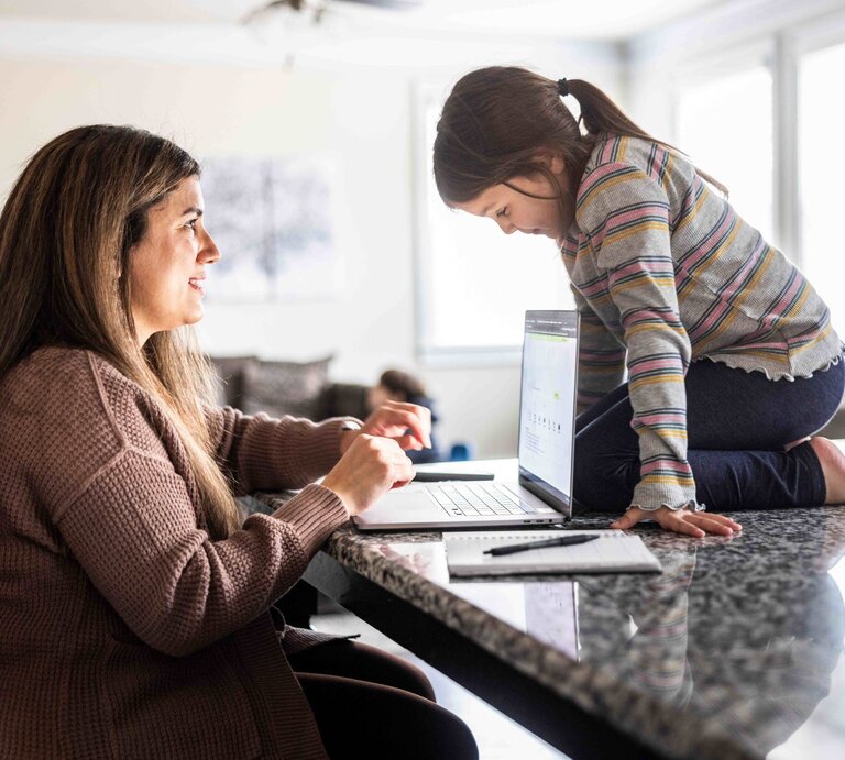 Das Bild zeigt eine am Küchentresen sitzende Frau vor ihrem Laptop. Auf dem Tresen kniet ein kleines Mädchen, das auf ihren Laptop schaut. | © Foto: Momo Productions | Getty Images 