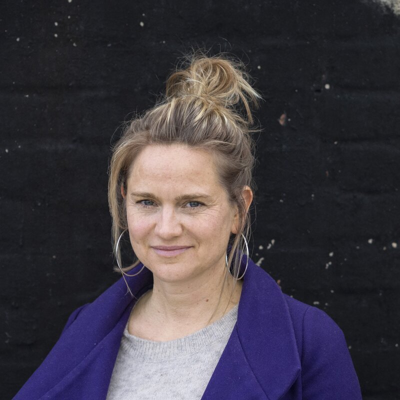 Autorin Eva Lohmann, blonder Zopf, oben am Kopf zusammengebunden, blickt vor schwarzem Hintergrund direkt in die Kamera.