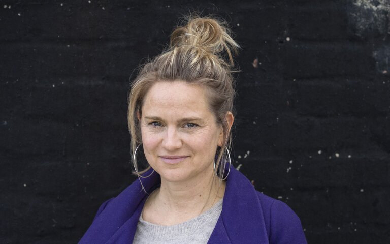 Autorin Eva Lohmann, blonder Zopf, oben am Kopf zusammengebunden, blickt vor schwarzem Hintergrund direkt in die Kamera.