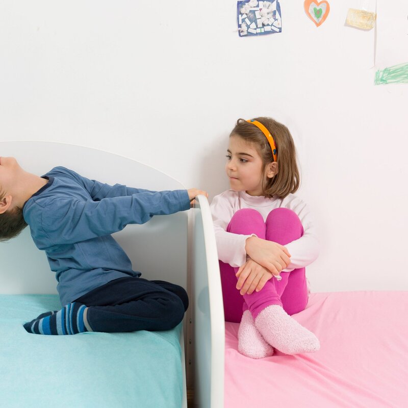 Man sieht die Enden von zwei Betten, die aneinandergereiht sind. Das linke Bett hat ein blaues Polster und drauf sitzt ein Junge mit einem blauen Pulli, der sich am Bettende festhält und nach hinten lehnt. Auf dem rechten Bett mit einem pinken Bezug sitzt ein Mädchen mit einer pinken Hose. Sie sitzt mit herangezogenen Knien an die hintere Wand gelehnt und schaut den Jungen an. | © Foto: bokan76 | Getty Images