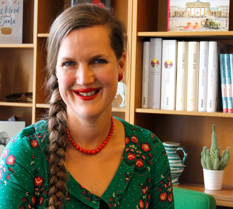 Frau in grünem Shirt blickt unmittelbar und lächelnd in die Kamera. Im Hintergrund steht ein Bücherregal mit vorwiegend weißen Buchrücken. | © Foto: Piper Verlag