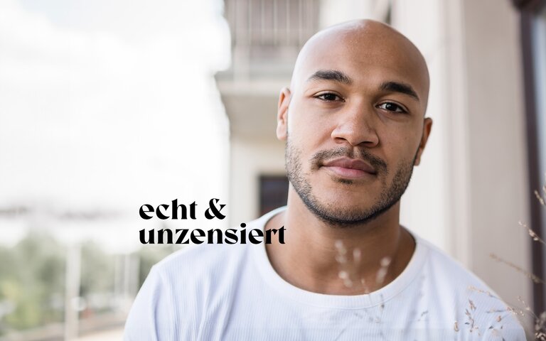 Schauspieler Malick Bauer steht auf einem Balkon und trägt ein weißes T-Shirt. | © Lucas Lentes